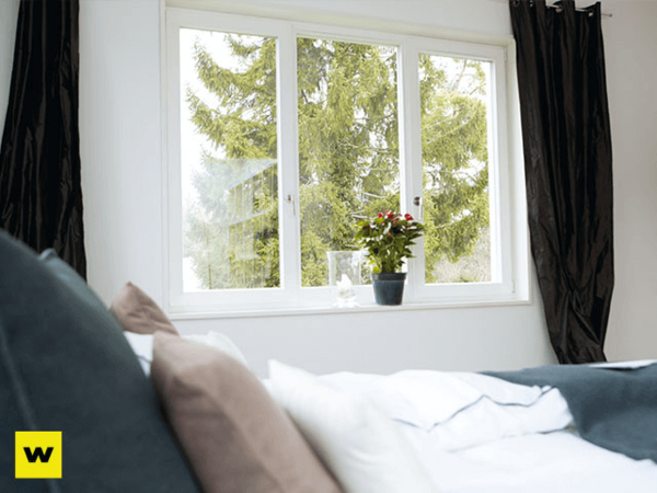 Helles Schlafzimmer mit Doppelbett, großen Fenstern und Ausblick auf grüne Bäume.