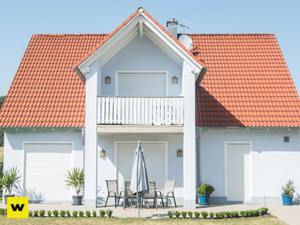 Modernes Einfamilienhaus mit rotem Dach, Terrasse und geschlossenen Rollläden im Sonnenschein.